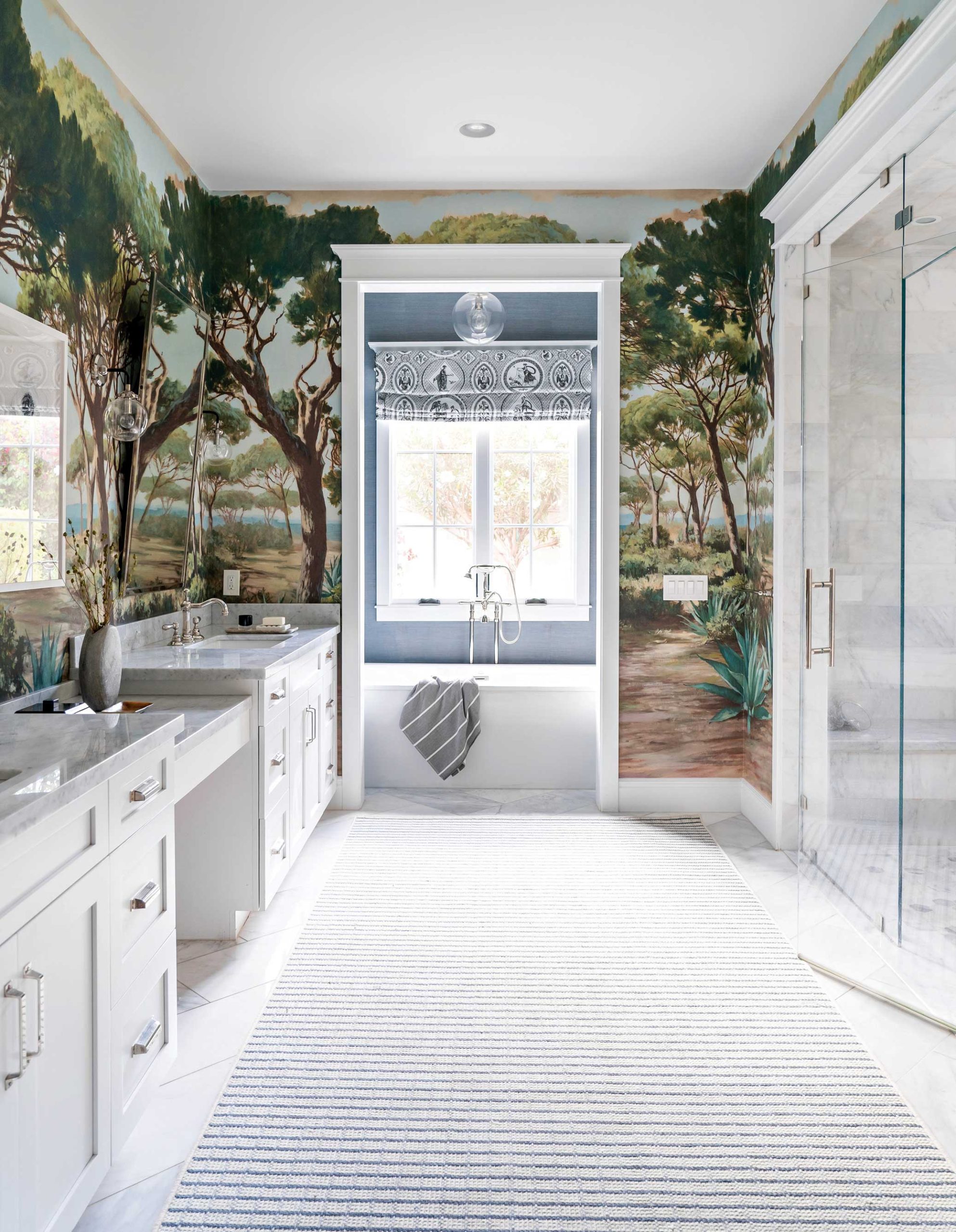 A landscape mural wallcovering transforms the previously white primary bath into a restful forest. The Roman shade over the tub is a toile fabric, upon which classical figures sport new technology, such as selfie sticks. A landscape mural wallcovering transforms the previously white primary bath into a restful forest. The Roman shade over the tub is a toile fabric, upon which classical figures sport new technology, such as selfie sticks.